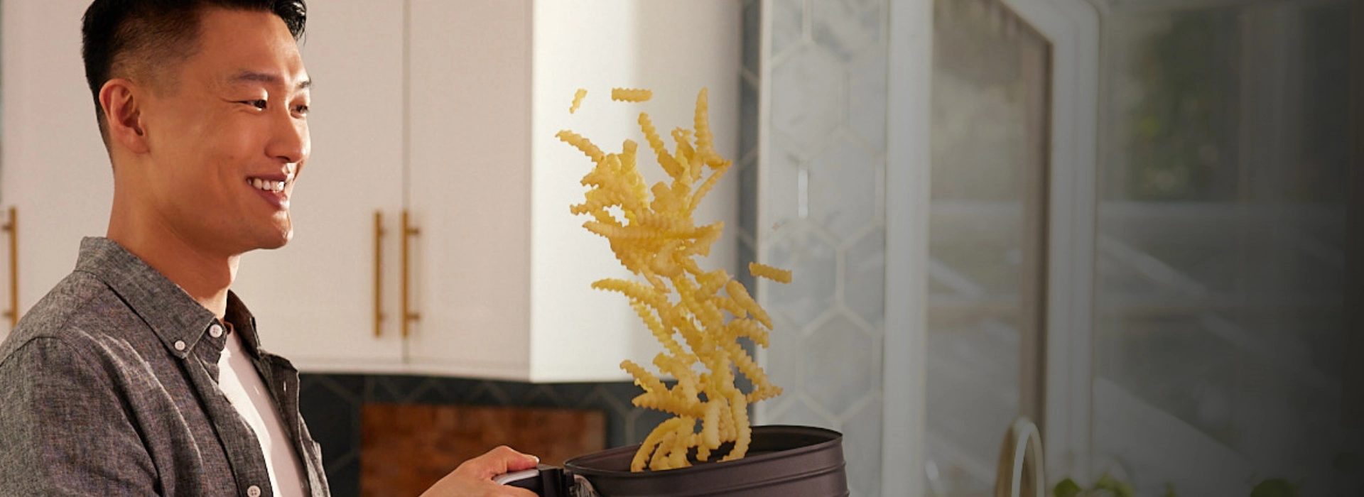 Smiling man in a modern kitchen with white cabinets and tiled walls tossing McCain crinkle-cut fries from a black pot